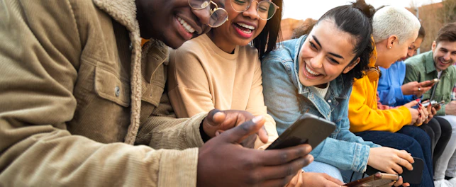 Happy multiracial teen college students having fun using mobile phone together in campus outdoors