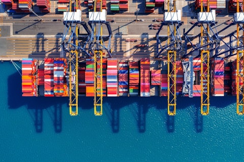 Aerial view of a busy shipping port with cranes loading and unloading colourful cargo containers along the waterfront.