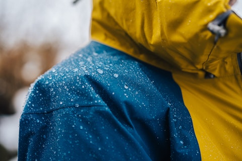 Close-up of a waterproof jacket with yellow and blue panels, covered in water droplets, indicating it is being worn in wet conditions