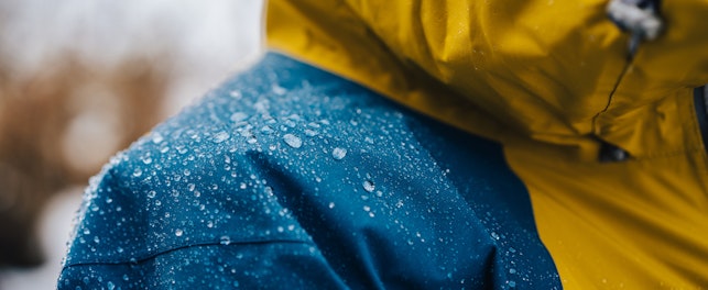 Close-up of a waterproof jacket with yellow and blue panels, covered in water droplets, indicating it is being worn in wet conditions