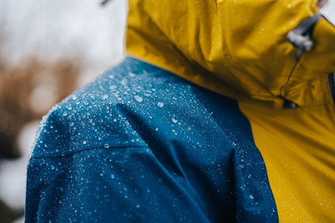 Close-up of a waterproof jacket with yellow and blue panels, covered in water droplets, indicating it is being worn in wet conditions