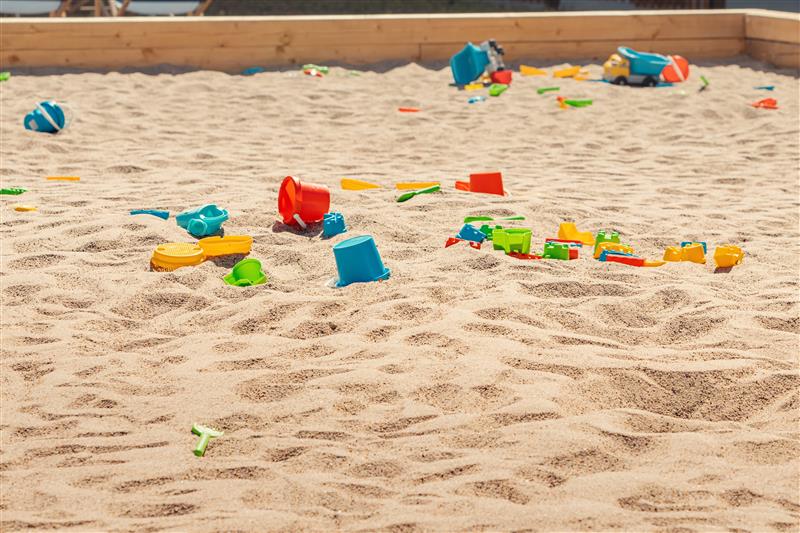 Children’s sandpit with colourful plastic toys and moulds scattered across the sand, including buckets, trucks, and small play tools.