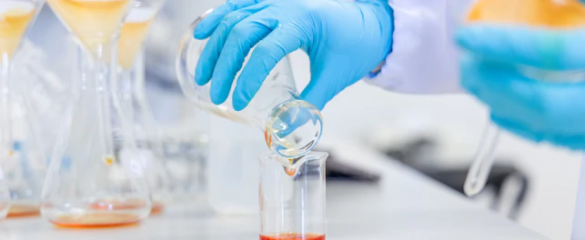 Hands pouring an orange chemical solution into a beaker