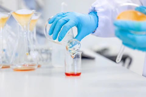 Hands pouring an orange chemical solution into a beaker