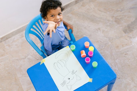 Happy Colombian Boy Having Fun Painting a Cat Drawing With Colorful Finger Paints at His Blue Table