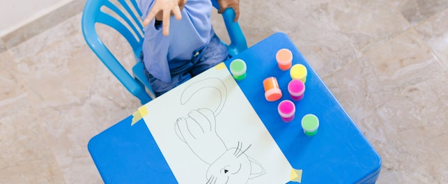 Happy Colombian Boy Having Fun Painting a Cat Drawing With Colorful Finger Paints at His Blue Table