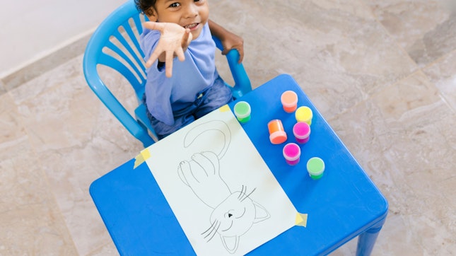 Happy Colombian Boy Having Fun Painting a Cat Drawing With Colorful Finger Paints at His Blue Table