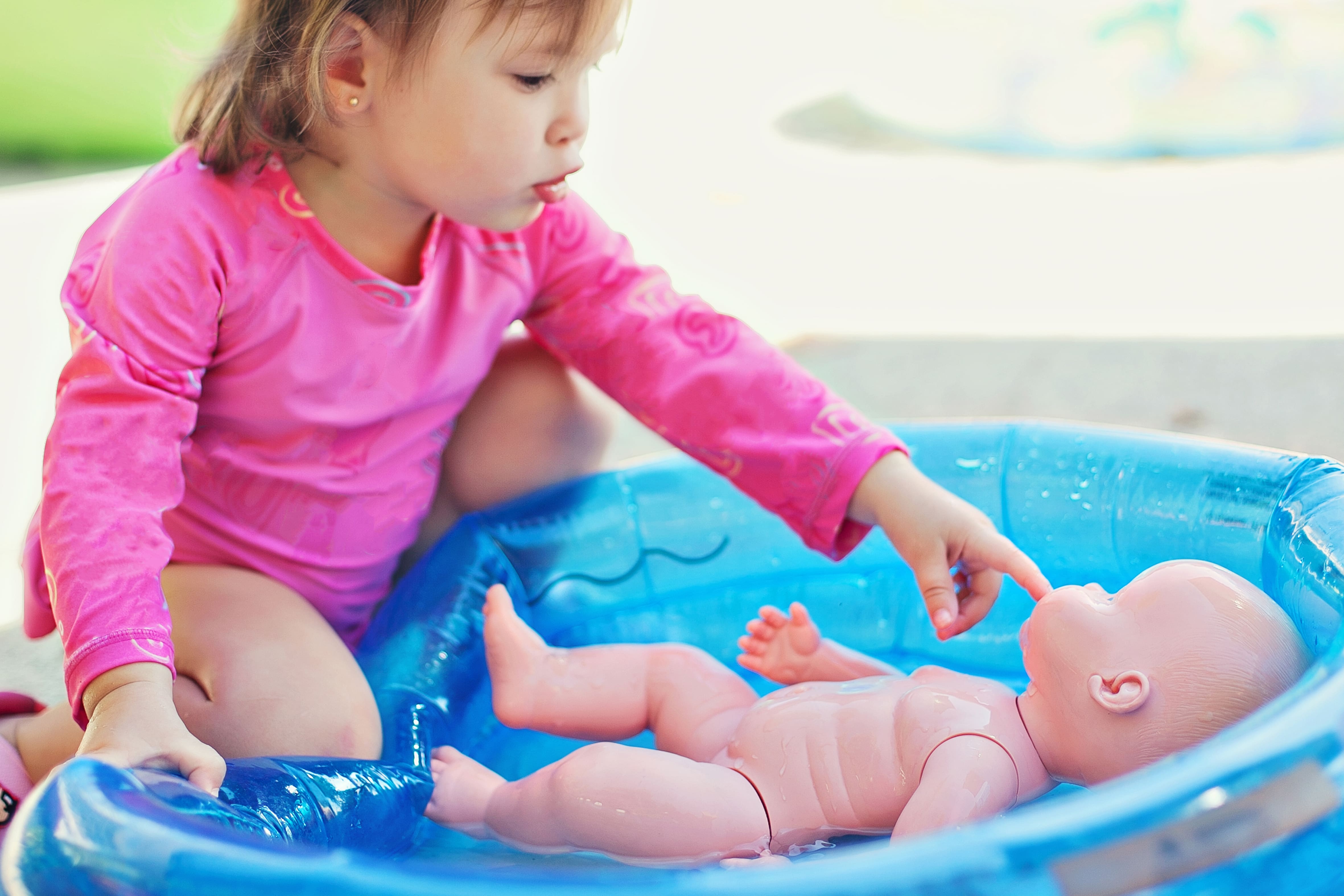 Une petite fille en tenue rose jouant avec une poupée dans une petite piscine gonflable.