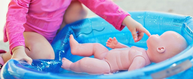 Une petite fille en tenue rose jouant avec une poupée dans une petite piscine gonflable.