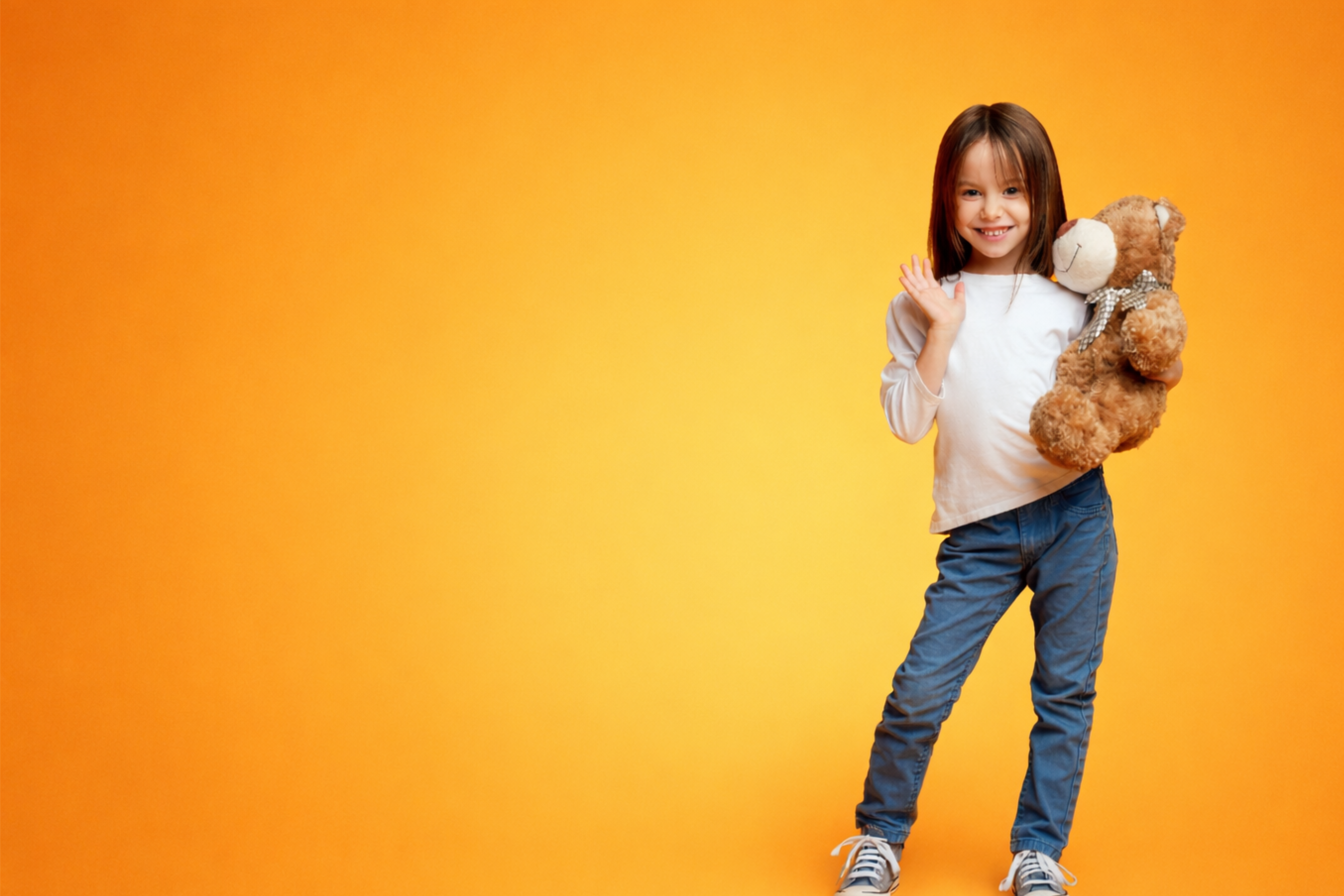Child standing against an orange background holding a brown teddy bear