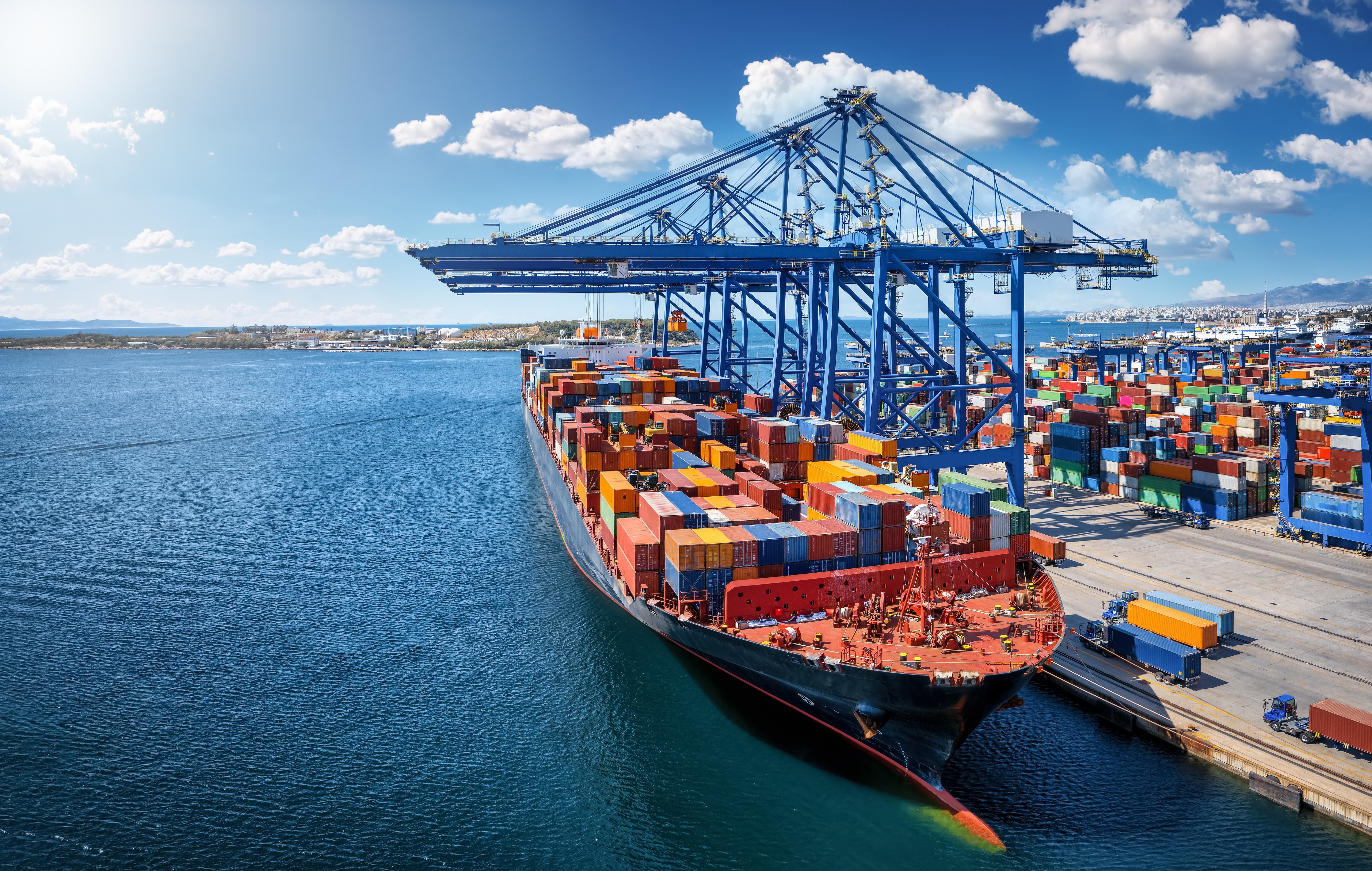 Large container ship docked at a busy seaport, with towering cranes loading and unloading stacks of colorful shipping containers along the quay.