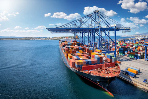 Large container ship docked at a busy seaport, with towering cranes loading and unloading stacks of colorful shipping containers along the quay.