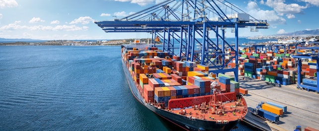 Large container ship docked at a busy seaport, with towering cranes loading and unloading stacks of colorful shipping containers along the quay.