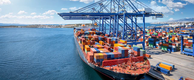 Large container ship docked at a busy seaport, with towering cranes loading and unloading stacks of colorful shipping containers along the quay.
