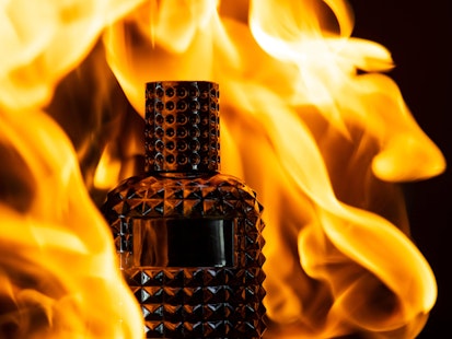 bouteille de parfum posé sur de la terre et entourée par des flammes sur fond noir 