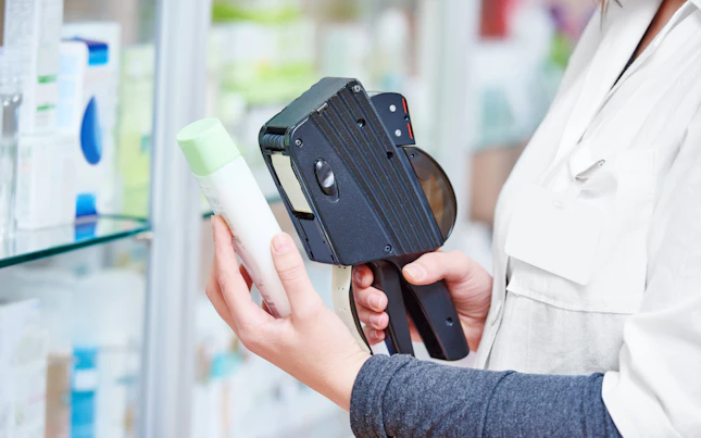 Hand van vrouwelijke apotheker die een etiketteerpistool gebruikt om een prijskaartje van een geneesmiddel in de drogisterij te plakken