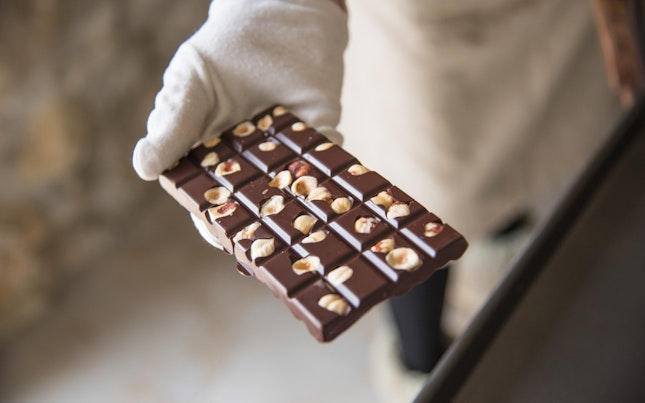 White Gloved Hand Holding a Hand Crafted Dark Chocolate and Hazelnut Bar Against Light Background Of