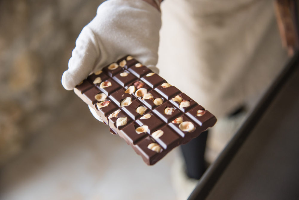 Witte gehandschoende hand houdt een handgemaakte reep pure chocolade en hazelnoot tegen een lichte achtergrond van