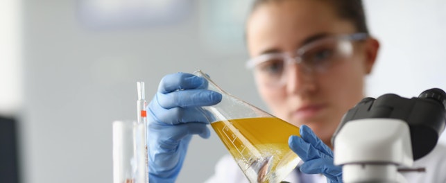 Scientifique en laboratoire analysant un liquide jaune dans un erlenmeyer, entourée de tubes à essai et d'un microscope.