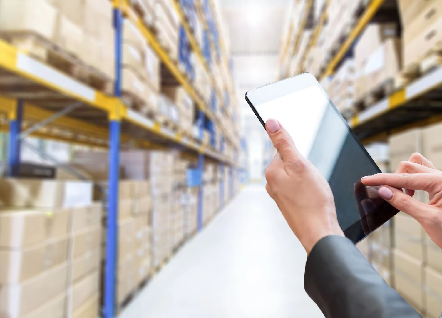 Horizontal color image of female hands holding a digital tablet in a corridor of futuristic distribution warehouse. Ordering online from a modern warehouse on a touchscreen tablet computer. Large distribution storage in background with racks full of packages, boxes, pallets, crates ready to be delivered. Logistics, freight, shipping, receiving.