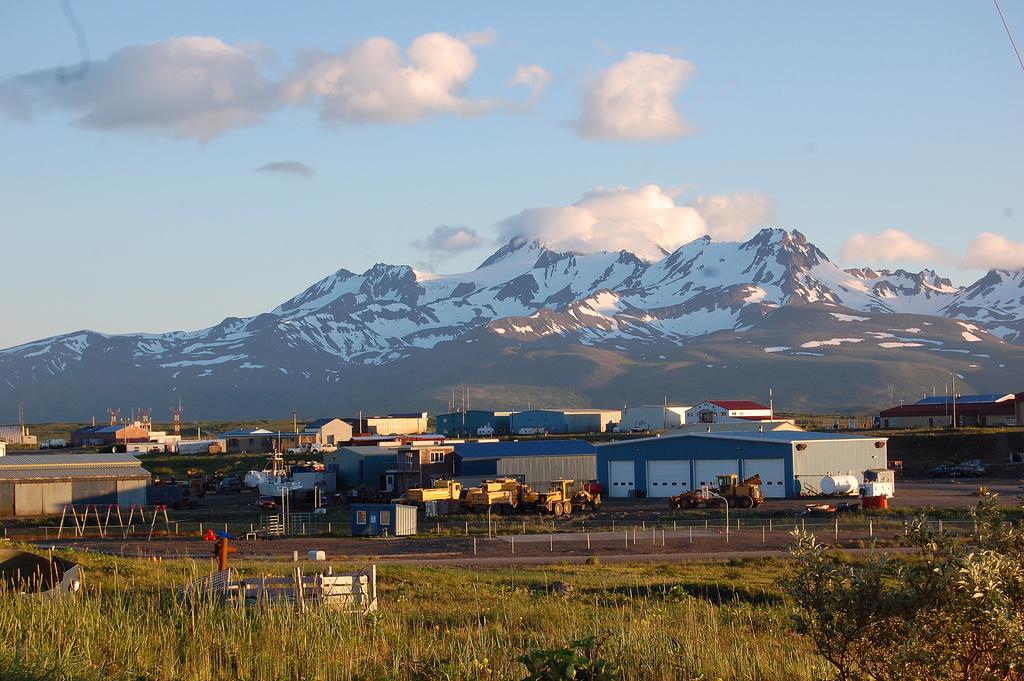 Industrial buildings and equipment beneath snow-covered mountains in Alaska under a blue sky with scattered clouds.