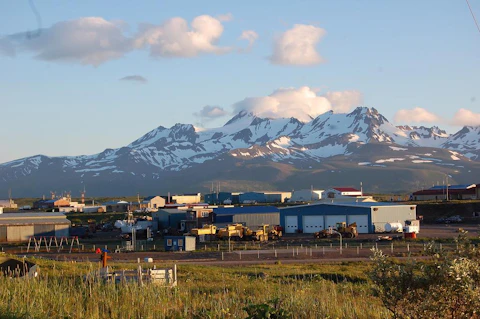 Industrial buildings and equipment beneath snow-covered mountains in Alaska under a blue sky with scattered clouds.