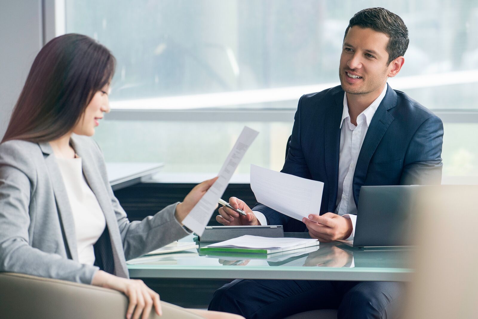 Two professionals review printed documents during a business meeting at a glass conference table with a laptop and paperwork nearby.