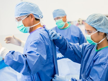 Female Surgical Technician or Nurse is Helping Lead Surgeon Put on Sterile Gown and Gloves