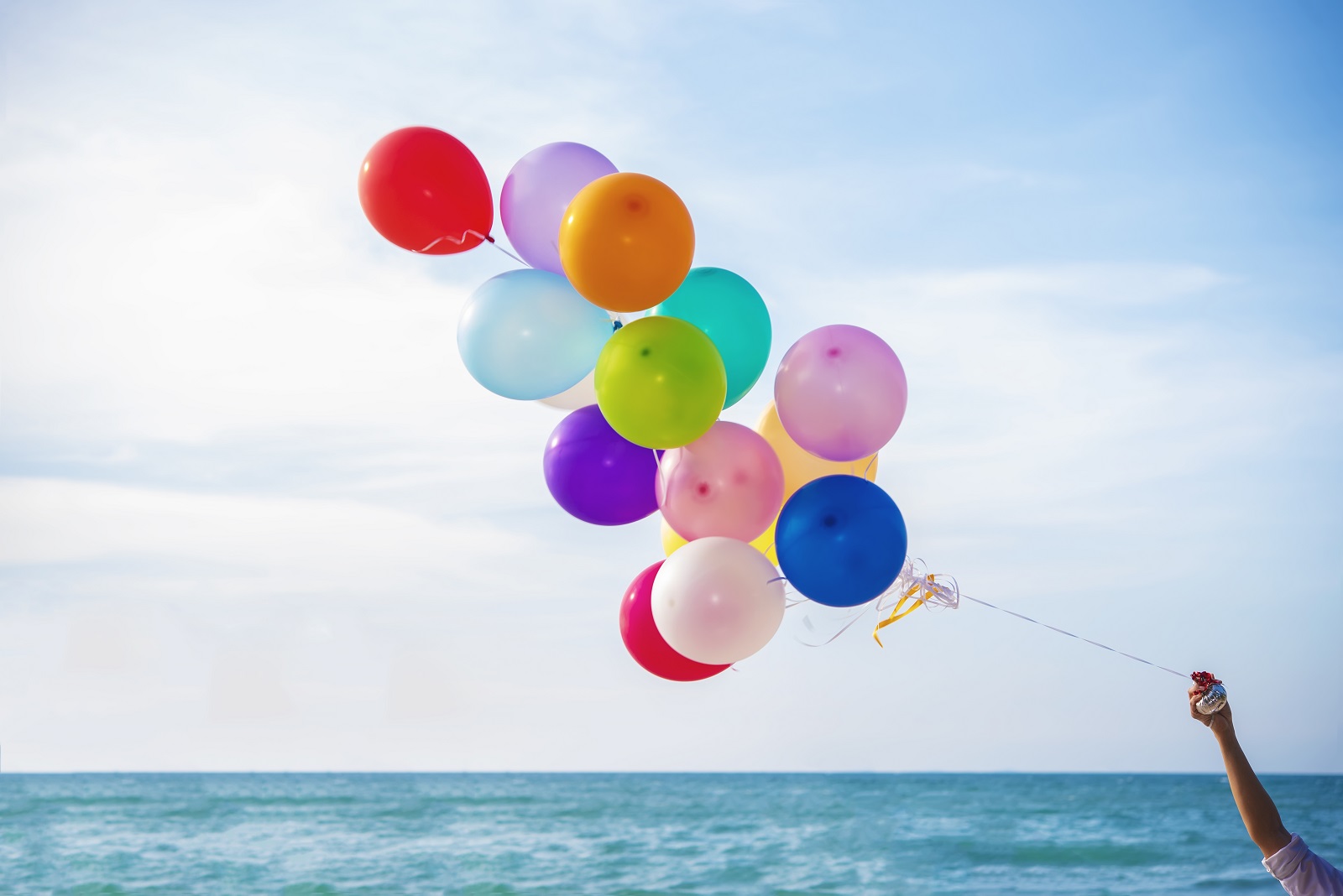 Dłoń trzymająca kolorowe balony, w tle widoczne niebo i morze.