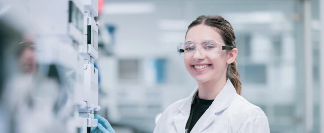 Uśmiechnięta pracownica SGS w fartuchu, z rękawiczkami i okularami ochronnymi, stojąca przy sprzęcie laboratoryjnym.